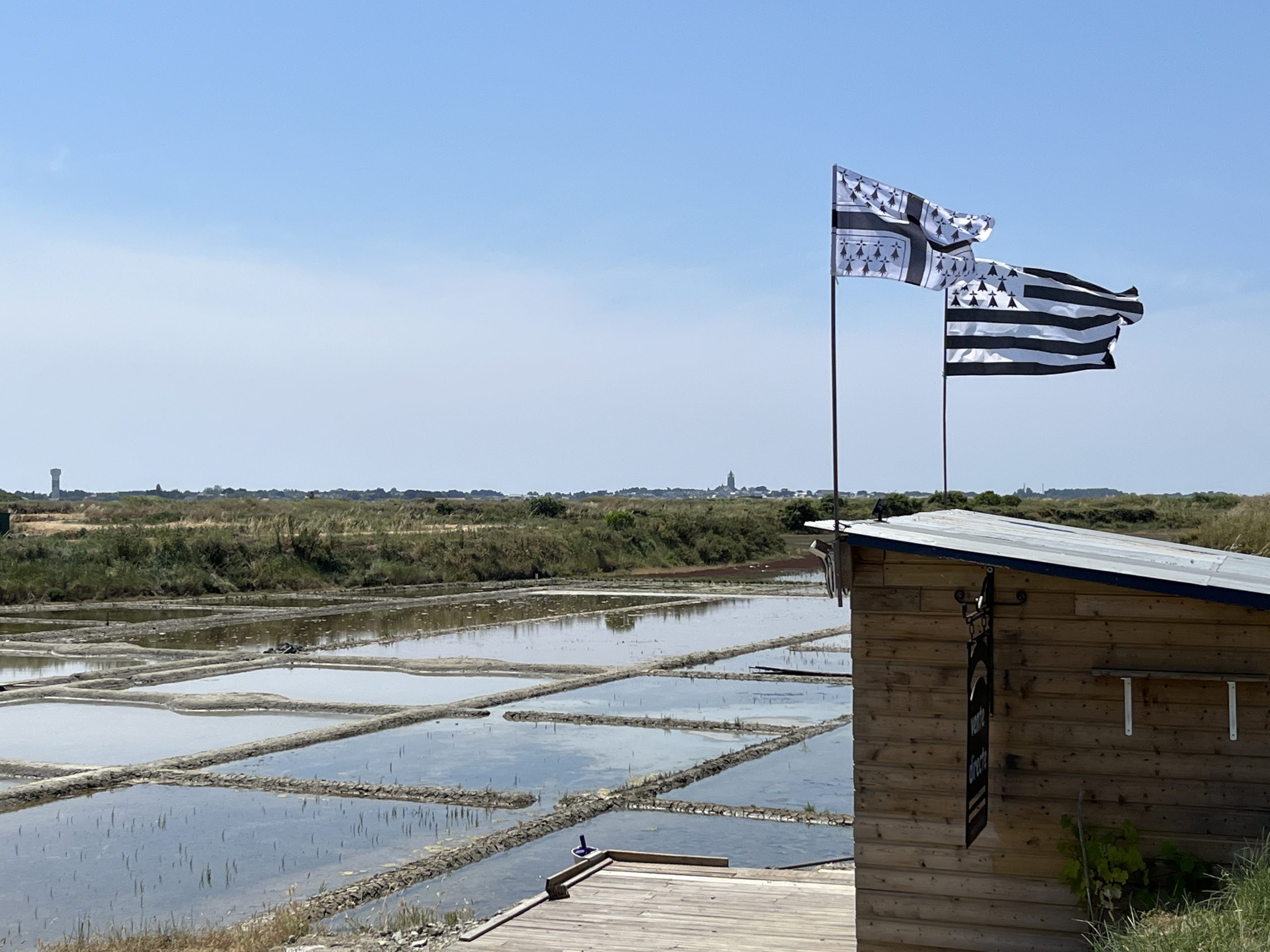 Vue des œillets des marais salants avec une petite cabane en bois au centre et deux drapeaux bretons flottant au-dessus.