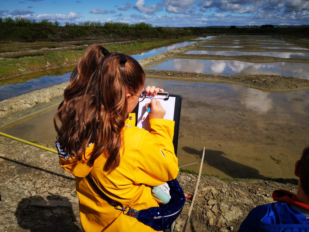 Une enfant devant un œillet des marais salants qui écrit sur un bloc-notes.