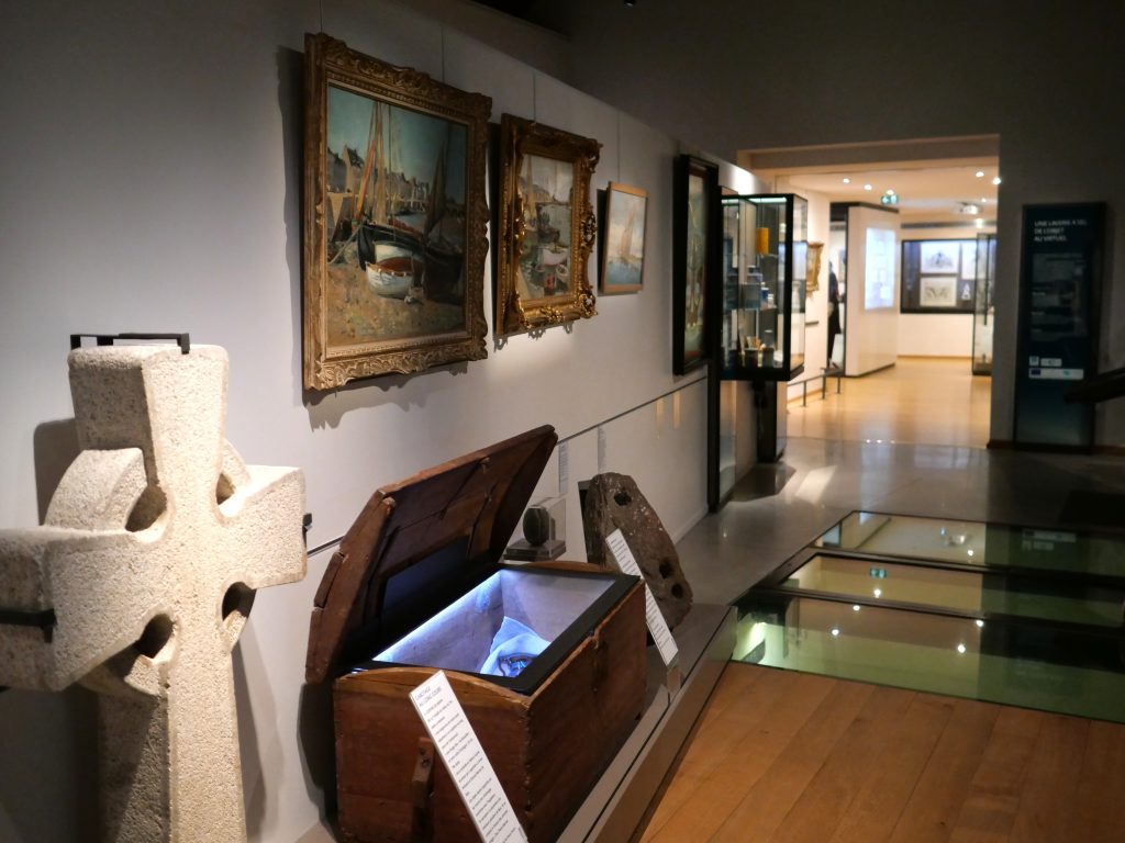 Quatre peintures du port du Croisic accrochées au mur, avec un coffre en bois et une croix en pierre posés au sol.